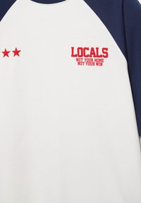T-shirt raglan blanc et marine avec le texte rouge "LOCALS NOT YOUR HOME NOT YOUR WIN" et deux étoiles rouges sur la poitrine.