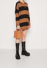 Abito maglione lavorato a maglia con strisce nere e marroni, colletto con mezza zip, abbinato a una piccola borsa a tracolla marrone testurizzata e stivali neri con lacci alti fino al ginocchio.