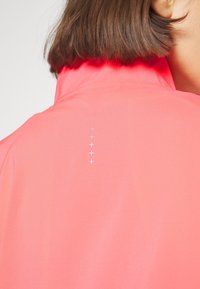 Włosy brązowe do ramion, pod jasnoróżową kurtką z wysokim kołnierzem, na plecach pięć małych odblaskowych krzyżyków w pionowej linii.