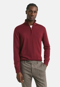 Junger Mann mit geflochtenem Haar, der einen weinroten Pullover mit Reißverschluss oben und graue Herringbone-Hosen trägt, steht mit einer Hand in der Tasche.