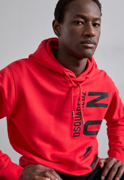 Haut à capuche rouge avec un cordon, présentant un grand design graphique noir sur le devant. Fabriqué en tissu doux avec une coupe décontractée.
