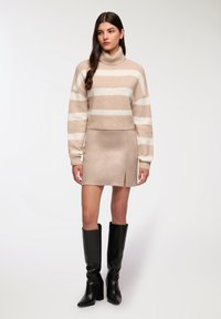 Maglione lavorato a maglia a righe crema e beige, mini gonna aderente in suede beige con spacco laterale, abbinato a stivali neri con tacco alti fino al ginocchio.