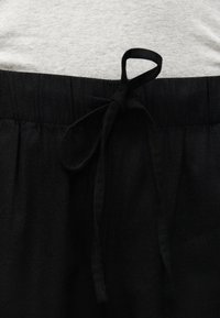 Λαστιχένια μέση μαύρου παντελονιού με δεμένη υφασμάτινη κορδονάκι πάνω σε φόντο από ελαφρώς γκρι ύφασμα πουκάμισου.