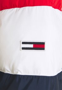 Piros, fehér és sötétkék szélvédődzseki, texturált anyagból, vörös, fehér és fekete hímzett logóval egy fehér panelen, téglalap alakban.