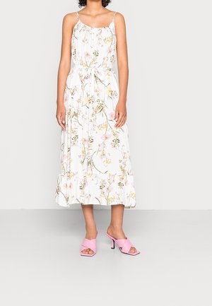 Frau trägt ein weißes ärmelloses Midikleid mit pinkem und gelbem Blumenmuster und pinken Sandalen mit Absatz, steht vor einem einfarbigen Hintergrund.