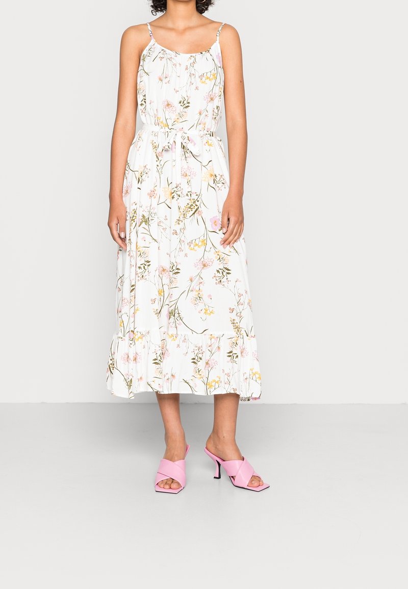 Femme portant une robe midi blanche sans manches avec un imprimé floral rose et jaune et des sandales à talons roses, debout devant un fond uni.