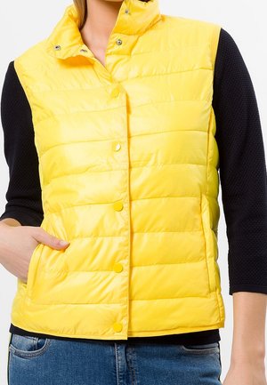 Gilet isolant jaune avec un col haut, des boutons-pression et des coutures horizontales. Porté sur un pull noir texturé. Coupe décontractée.