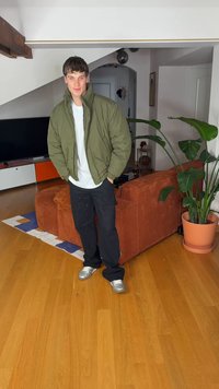 Zöld pufi dzseki, fehér póló, fekete nadrág, ezüst tornacipő. Fa padlón áll barna kordbársony kanapé és beltéri növény mellett.