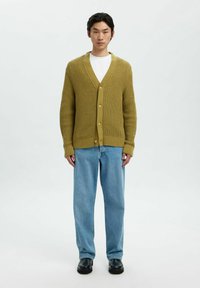 Man som bär en olivgrön strukturerad knäppkardigan, vit skjorta, lösa ljusblå jeans och svarta skor, står mot en helt vit bakgrund.