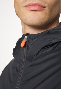 Schwarze Zip-Jacke mit hohem Kragen, ausgestattet mit einem metallischen Reißverschluss und einem orangen Ziehband. Glattes Material mit leichtem Textur.