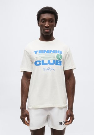 Férfi fehér "Tennis Club" feliratú pólóban és fehér "Borg" feliratú rövidnadrágban, egyszínű, világos háttér előtt áll.