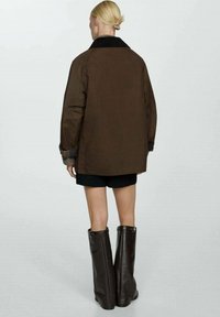 Brun oversized jakke med en sort krave, manchetter med ternet detalje, båret over sorte shorts og høje brune læderstøvler. Enked background.