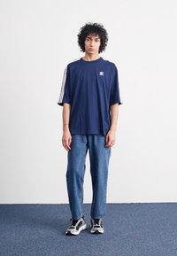Marineblå trænings t-shirt med hvide striber på de korte ærmer og et lille logo, parret med løstsiddende lyseblå denimjeans og sneakers.