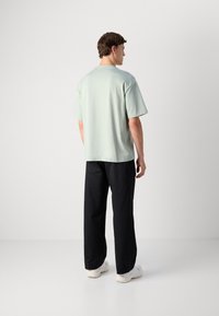 Moški obrnjen stran v svetlem zelenem oversized majici, črnih ohlapnih hlačah in belih supergah na enobarvni beli ozadju.