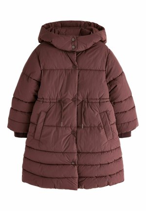 Unge pige med langt hår i to fletninger iført en lang bordeaux dunjakke, blå vidde jeans og hvide sneakers, stående foran en neutral baggrund.