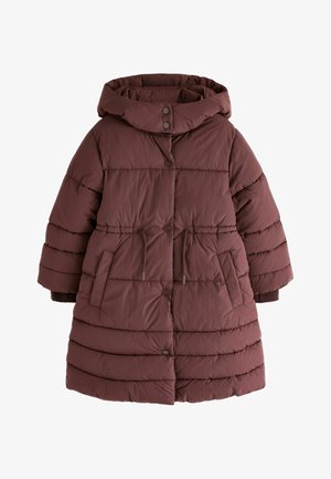 Unge pige med langt hår i to fletninger iført en lang bordeaux dunjakke, blå vidde jeans og hvide sneakers, stående foran en neutral baggrund.