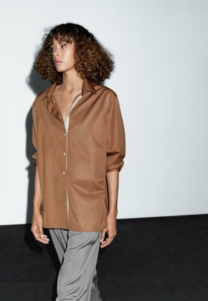 Frau mit lockigem Haar, die ein lockeres braunes Hemd mit Knöpfen und graue Hosen trägt, steht vor einem weißen und schwarzen Hintergrund.