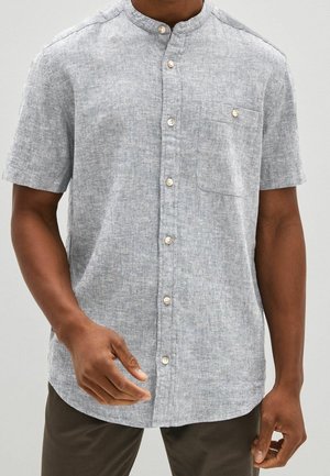 Homme portant une chemise en lin gris clair à manches courtes avec un col mandarin et des boutons sur le devant, associée à un pantalon sombre.
