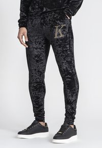 Zwarte fluwelen joggers met een slim fit, voorzien van een gouden sequin "K" accent op de zak. Gecombineerd met zwarte sneakers met witte zolen.