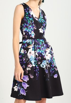 Černé šaty bez rukávů s výstřihem do V, rozšířenou sukní a výrazným modrým, fialovým a bílým květinovým potiskem, které nosí modelka.