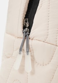 Sección de tela acolchada beige con una cremallera negra. Cuenta con un tirador de metal y dos tiradores de cremallera de silicona blanca texturizada. Detalle de costura negra visible.