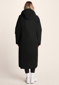 Woman wearing a long black hooded coat, black leggings, white socks, and white sneakers, standing facing away against a beige background.