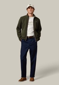 Olijfgroene fleecejas met ritssluiting aan de voorkant, wit T-shirt met logo, marineblauwe broek en bruine suède schoenen, gecombineerd met een witte pet.