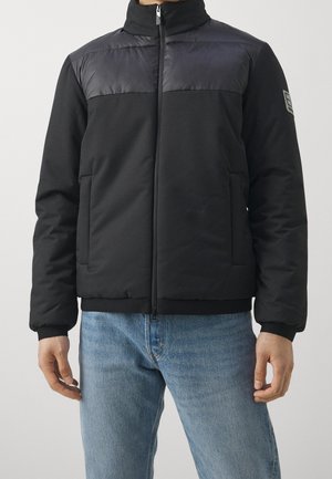 Mand iført en sort, polstret jakke med høj krave og lynlås foran, kombineret med blå jeans mod en ensfarvet baggrund.