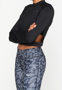 Person wearing a black cropped zip-up jacket and high-waisted gray patterned leggings with Puma logos, arms crossed over chest.