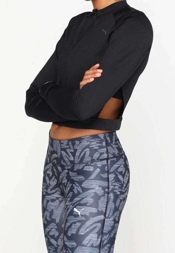 Person wearing a black cropped zip-up jacket and high-waisted gray patterned leggings with Puma logos, arms crossed over chest.