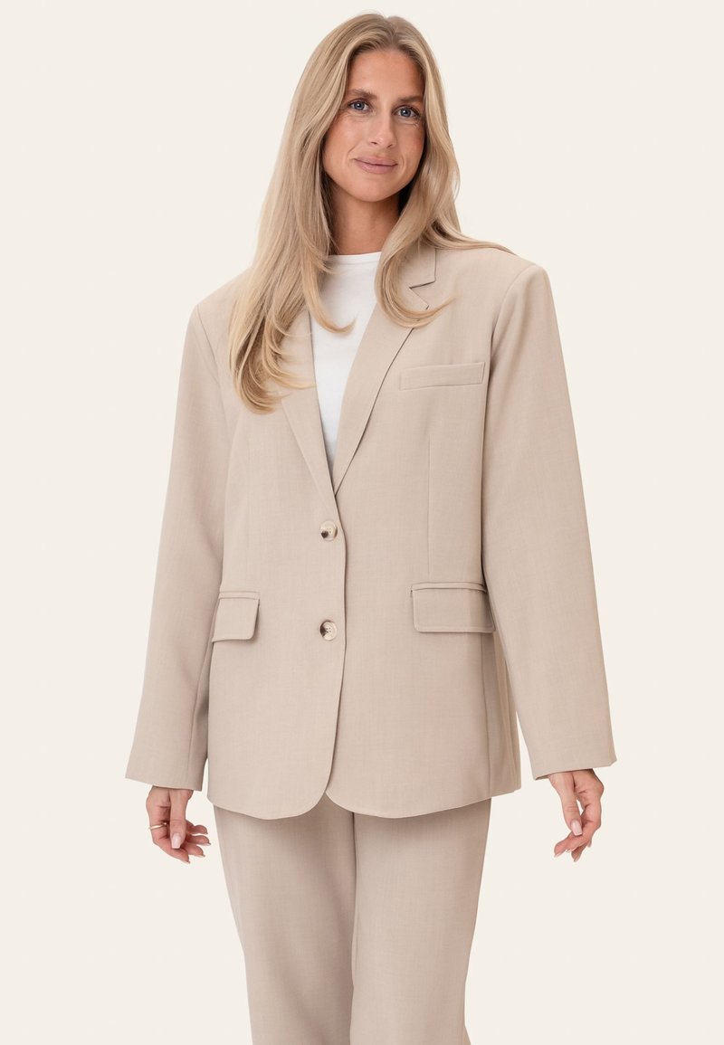 Femme aux longs cheveux blonds portant une veste de costume beige oversize et un pantalon assorti par-dessus un haut blanc, debout devant un fond uni.