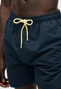 Άτομο με σκουρόχρωμο δέρμα που φορά σκούρο μπλε σορτς με κίτρινο κορδόνι δεμένο σε φιόγκο μπροστά, ορατό από τη μέση μέχρι το μέσο του μηρού.