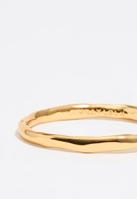 Pulsera de metal dorado en forma de bangle con un diseño retorcido y textura suave, que presenta un logo grabado en la superficie interna.