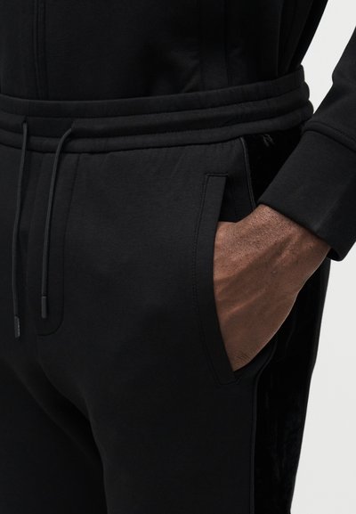 Svarte joggebukser med jevn tekstur og elastisk midjebånd. Har sidelommer og en fløyelsaktig detalj langs sømmene.