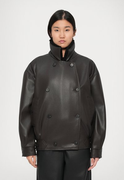 Young person with straight dark hair wears oversized dark brown leather jacket with high collar and button details, standing against plain background.