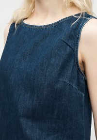 Γυναίκα που φοράει αμάνικη μπλε σκούρη denim μπλούζα με στρογγυλή λαιμόκοψη, οι ώμοι και το άνω μέρος του στήθους είναι ορατά.