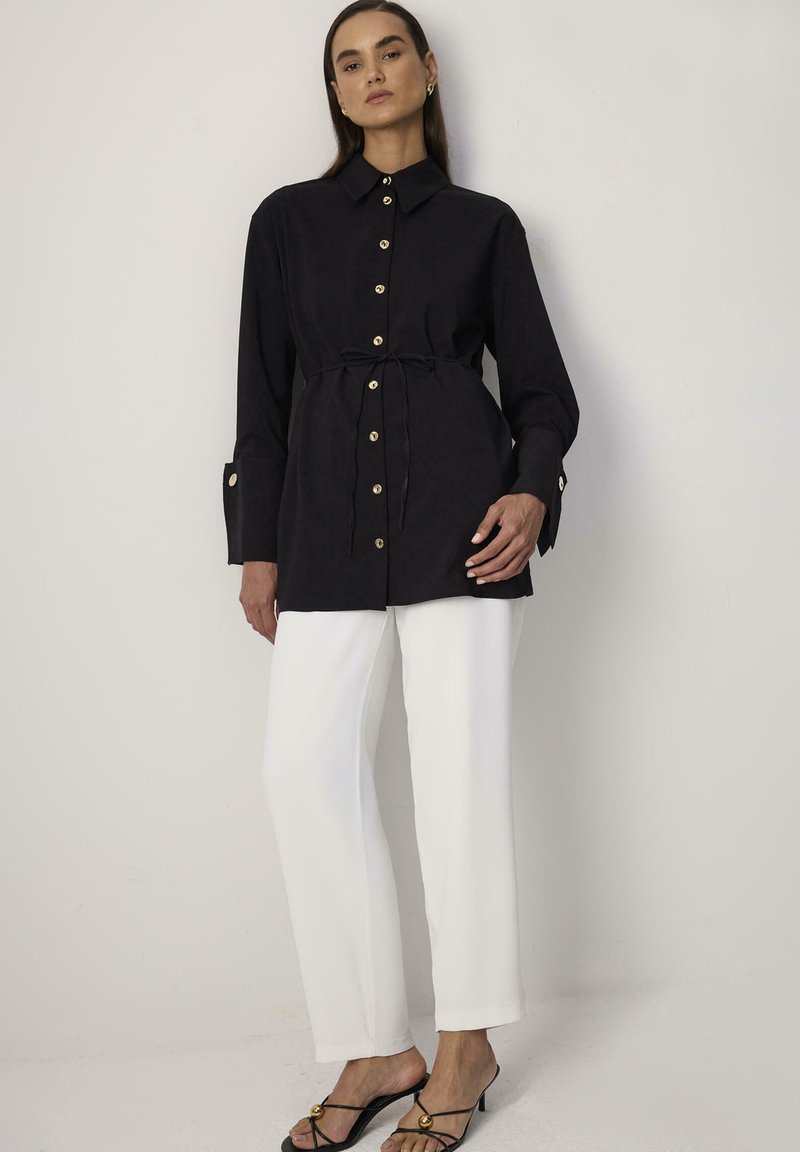 Camicia nera con bottoni, maniche lunghe, vita aderente, bottoni dorati e cintura, abbinata a pantaloni bianchi e semplici sandali neri.