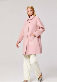 Manteau en laine rose avec un col rond, ornée de boutons dorés et d'une poche avant, assorti à un pantalon blanc et des chaussures argentées.