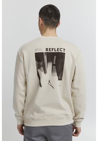 Sudadera de un beige claro con un diseño gráfico en la parte trasera que incluye la palabra "REFLECT" y una ilustración de un paisaje urbano. Puños y dobladillo acanalados.