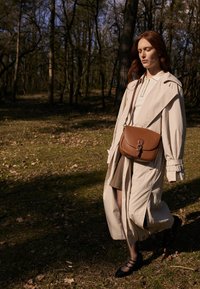 Lys beige trenchcoat over en grå kjole, brun læder skuldertaske, sorte sko, går på grønt græs med træer i baggrunden.