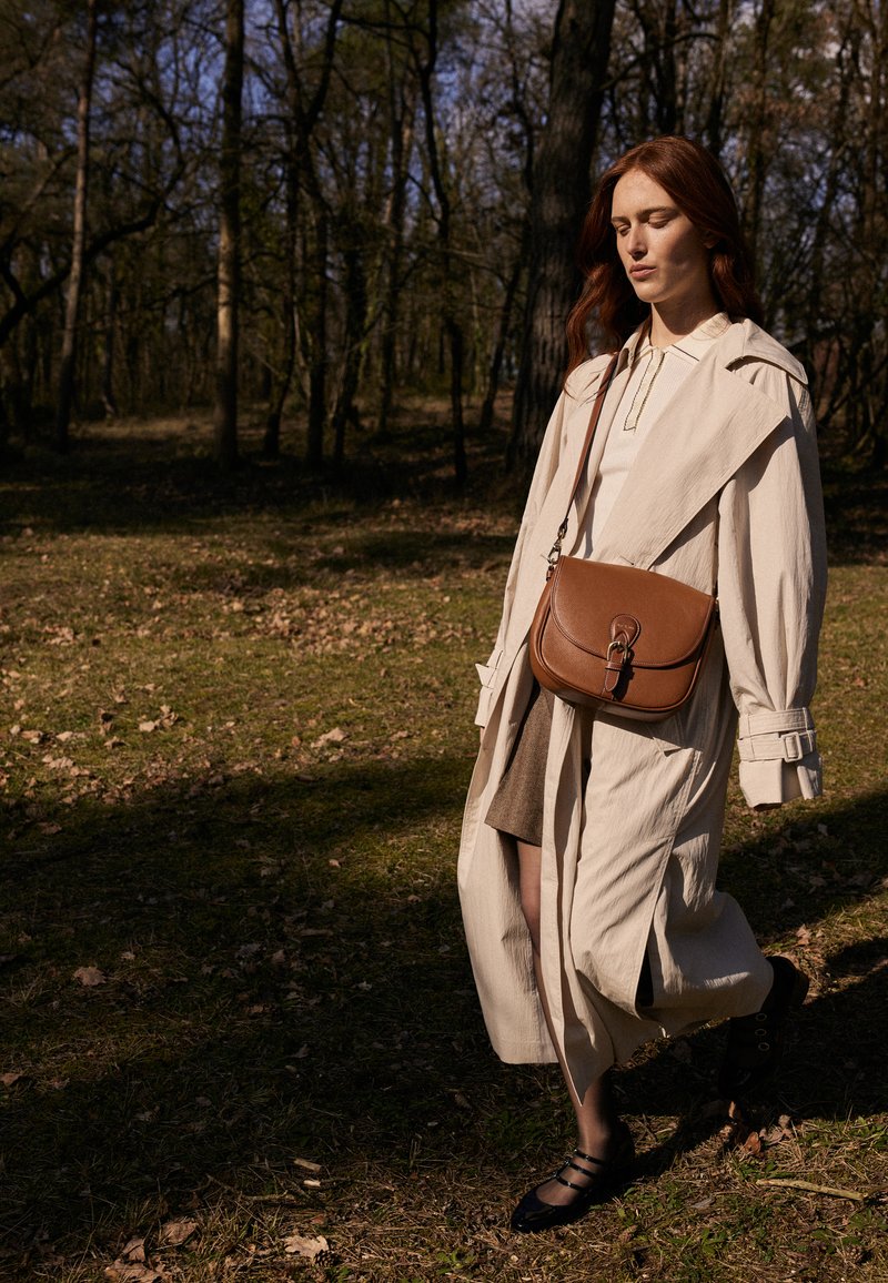 Lys beige trenchcoat over en grå kjole, brun læder skuldertaske, sorte sko, går på grønt græs med træer i baggrunden.