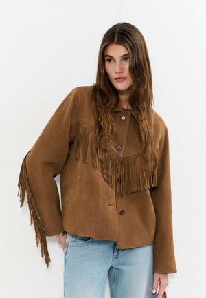 Jeune femme portant une veste en daim marron à franges avec des boutons et un jean bleu clair, posant devant un fond blanc.