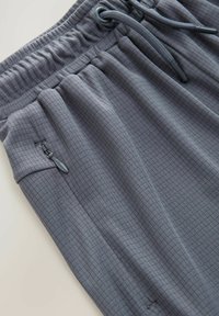 Pantalons de sport gris en tissu texturé, arborant un motif en quadrillage, avec une taille élastiquée et des poches avec fermeture éclair pour un rangement sécurisé.