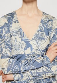 Femme portant un haut à manches longues à rayures florales bleues et beiges en V, les bras croisés sur la poitrine.