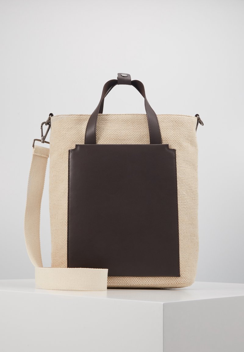 Sac en toile beige avec accents en cuir marron foncé, double poignée fixe et bandoulière amovible. Texture lisse et forme structurée.