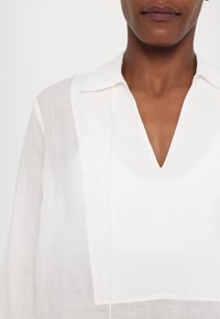Camicia bianca di lino con scollatura a V, colletto e vestibilità ampia, con dettagli di cucitura visibili sulla parte anteriore.
