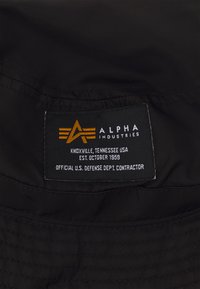Black nylon tag with white and orange lettering; features brand name "ALPHA INDUSTRIES," location, establishment date, and contractor details.
