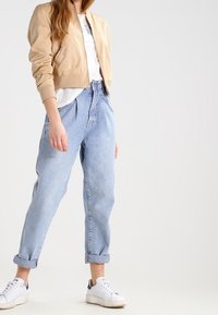 Žena v béžové bomber bundě, bílém tričku, světle modrých zastrčených džínách a bílých teniskách na jednobarevném pozadí.