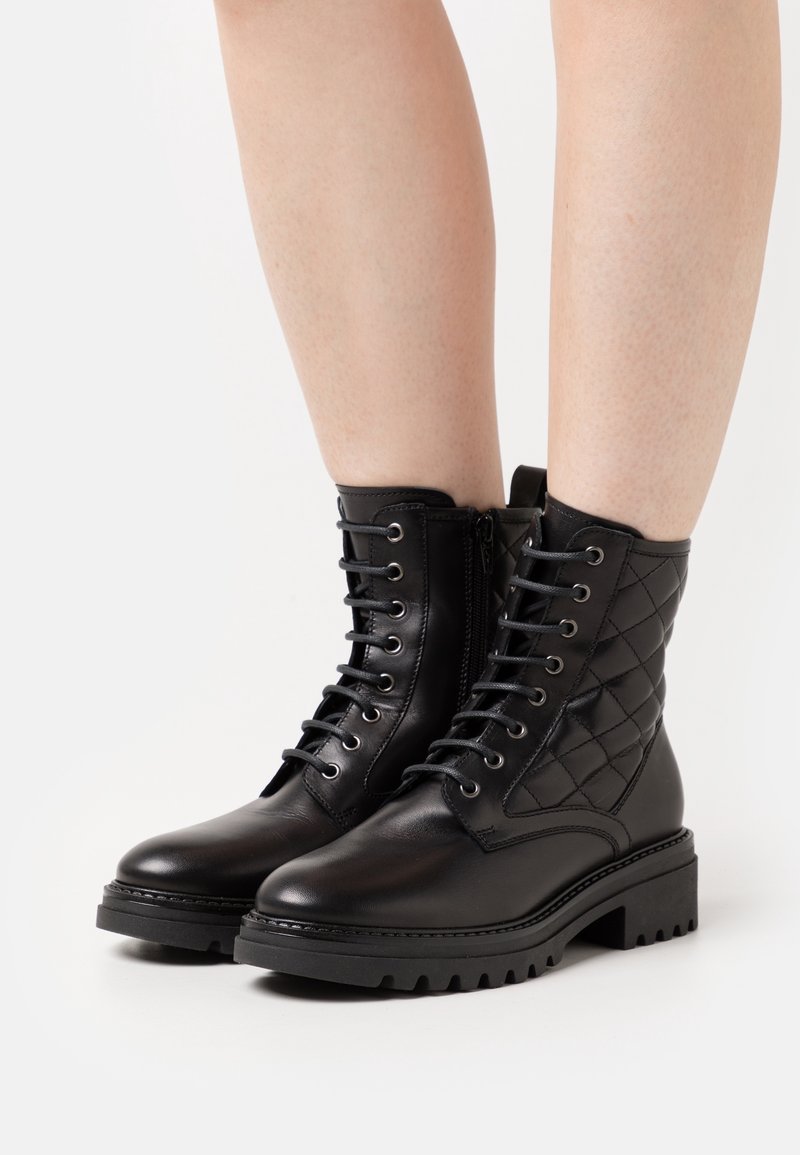 Bottines en cuir noir avec côtés matelassés, avant à lacets et semelles épaisses en caoutchouc, portées sur des jambes nues.
