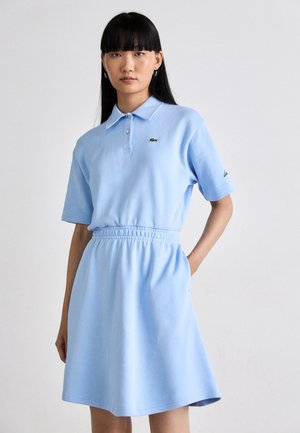 Vrouw met lang zwart haar die een lichtblauwe polo jurk met korte mouwen draagt, met een elastische taille en een geborduurd logo op de borst en mouw.